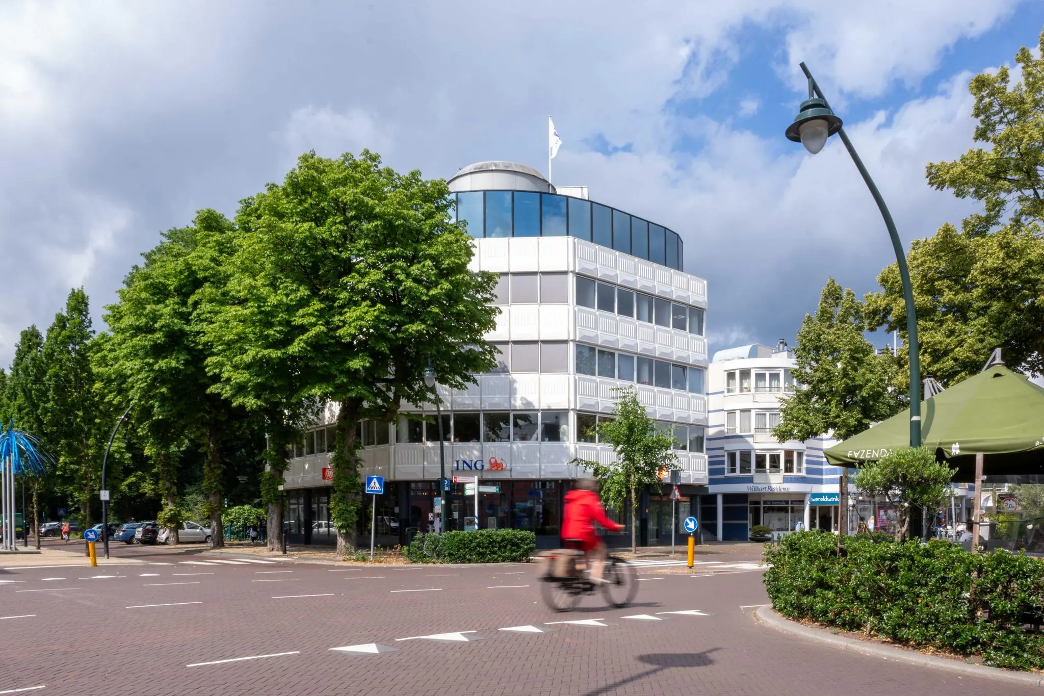 Straatbeeld van de 1e Hogeweg met een modern kantoorgebouw, een ING-filiaal en een fietser in een rood jack op de voorgrond.